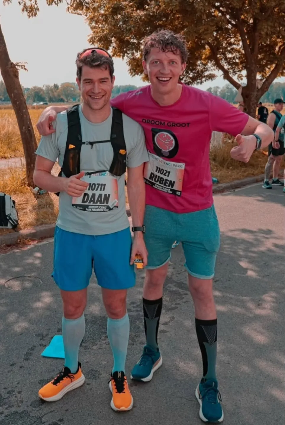 Ruben en zijn vriend Daan staan lachend naast elkaar bij de Utrecht Science Park Marathon, met startnummers op hun shirt. Ruben draagt een roze shirt met "Droom groot" erop, slaat een arm om Daan heen en steekt zijn duim op.