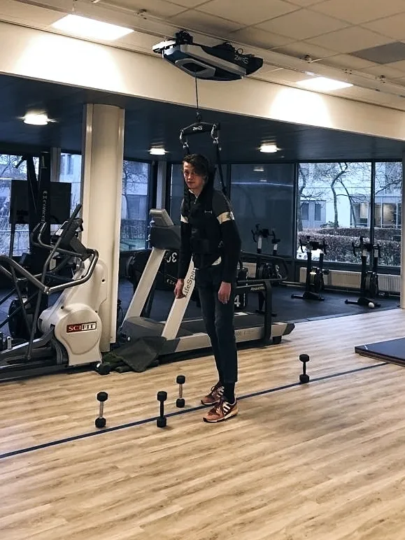 Ruben staat rechtop in de oefenzaal van het revalidatiecentrum, vastgemaakt aan een tuig dat aan een rail in het plafond hangt. Voor hem op de vloer staan kleine pionnen voor een looptraining.