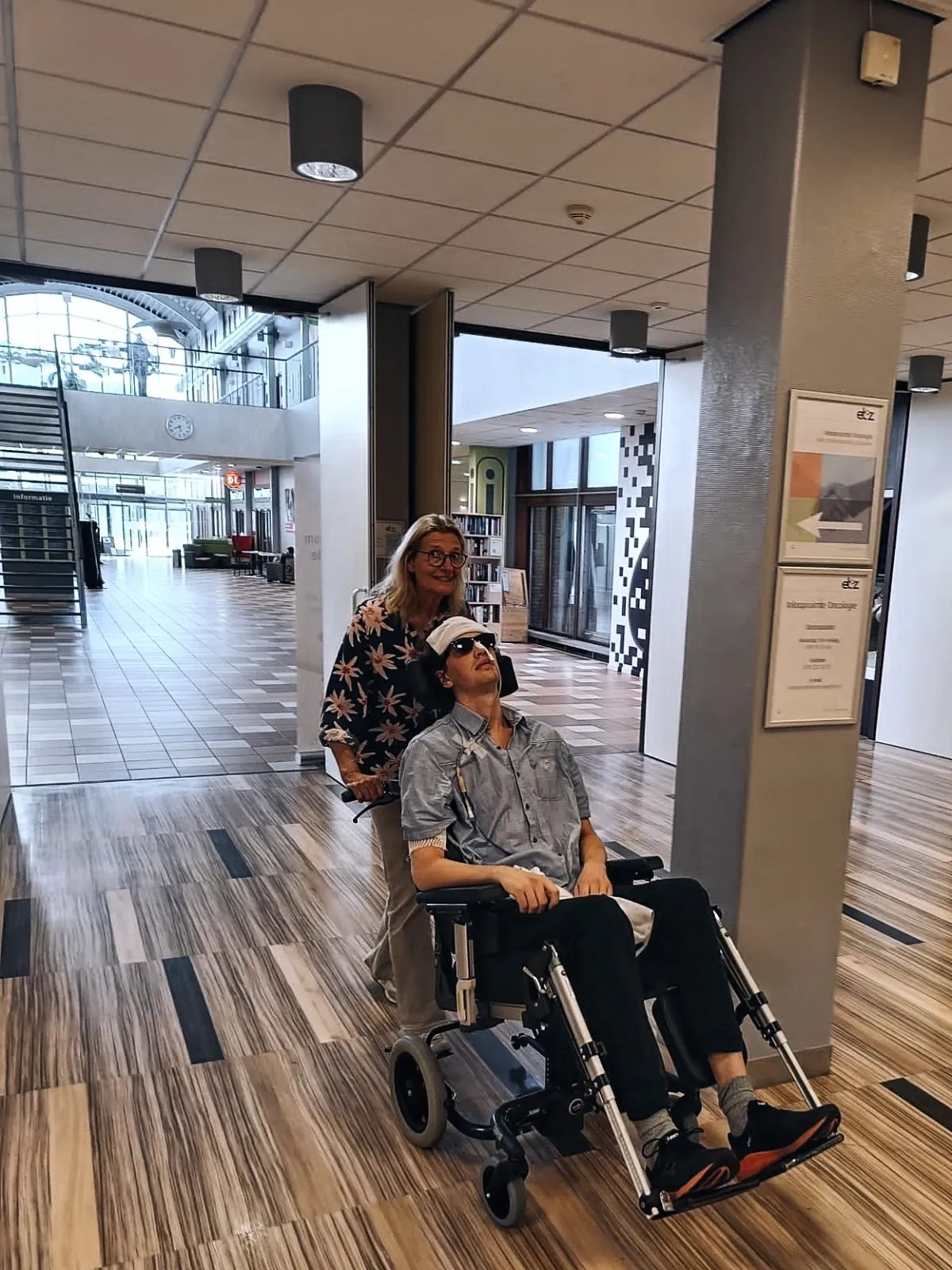 Ruben zit in een rolstoel met een witte pet op, in de lichte hal van het revalidatiecentrum. Een vrouw met een bloemetjesblouse duwt hem en glimlacht.