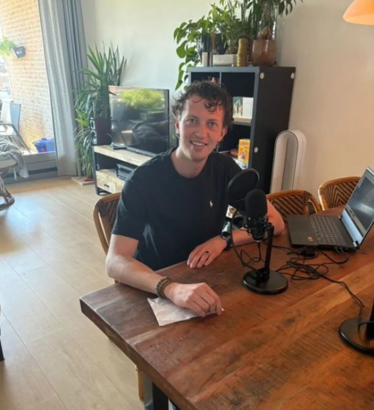 Ruben zit lachend aan een houten tafel in een woonkamer, met voor zich een podcastmicrofoon en naast zich een laptop, klaar om op te nemen.