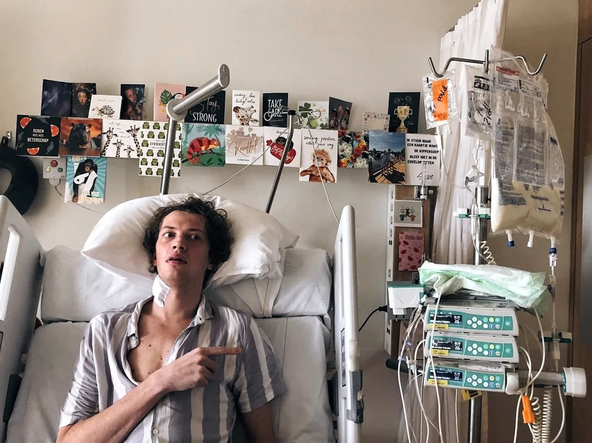 Ruben zit rechtop in een ziekenhuisbed, in een gestreept overhemd, en wijst met zijn vinger naar zijn borst. Achter hem hangt een rij kleurrijke kaarten aan de muur, rechts staan een infuuspaal en pompen.