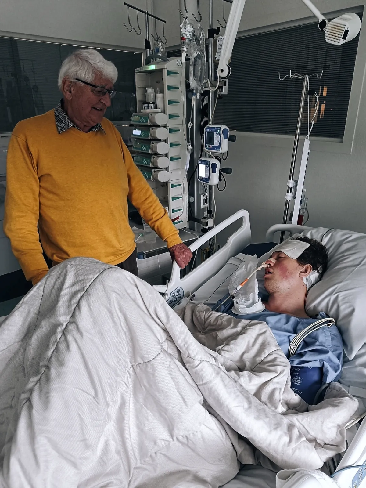 Ruben ligt in een ziekenhuisbed op de intensive care, met een verband op zijn voorhoofd en een slangetje in zijn neus. Naast het bed staat een oudere man in een gele trui die naar hem kijkt en zijn hand op de bedrand legt.