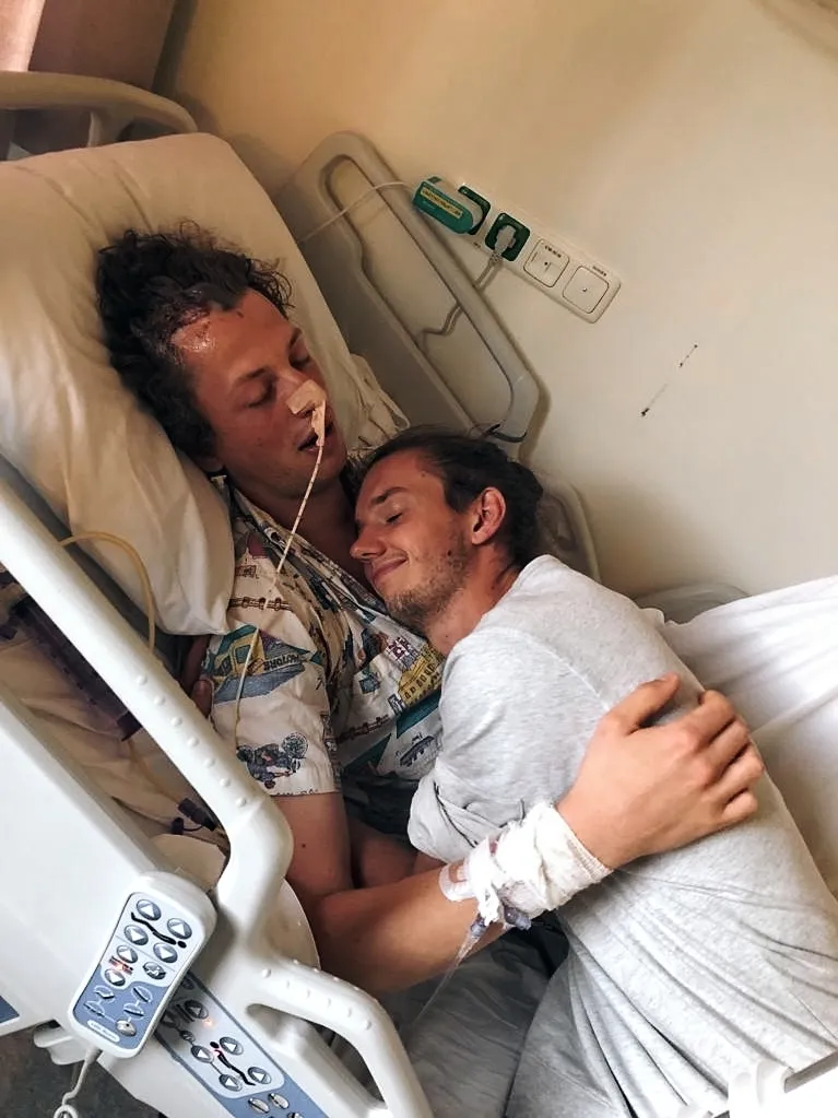 Ruben ligt met gesloten ogen in een ziekenhuisbed, met een slangetje in zijn neus. Een jonge man ligt naast hem op het bed en houdt hem stevig vast, met zijn hoofd tegen dat van Ruben.